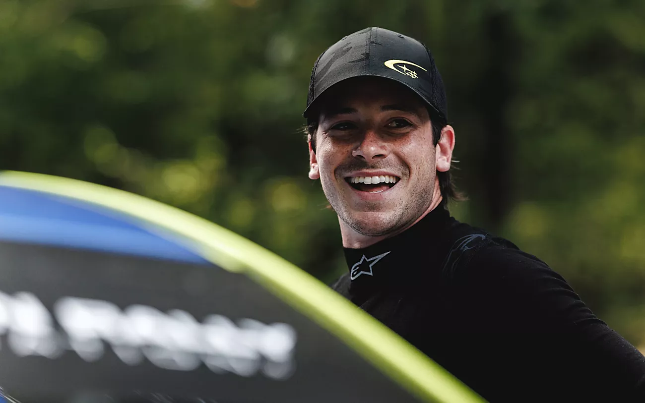 Conner Martell wearing a Subaru cap and race suit beside the rally car.
